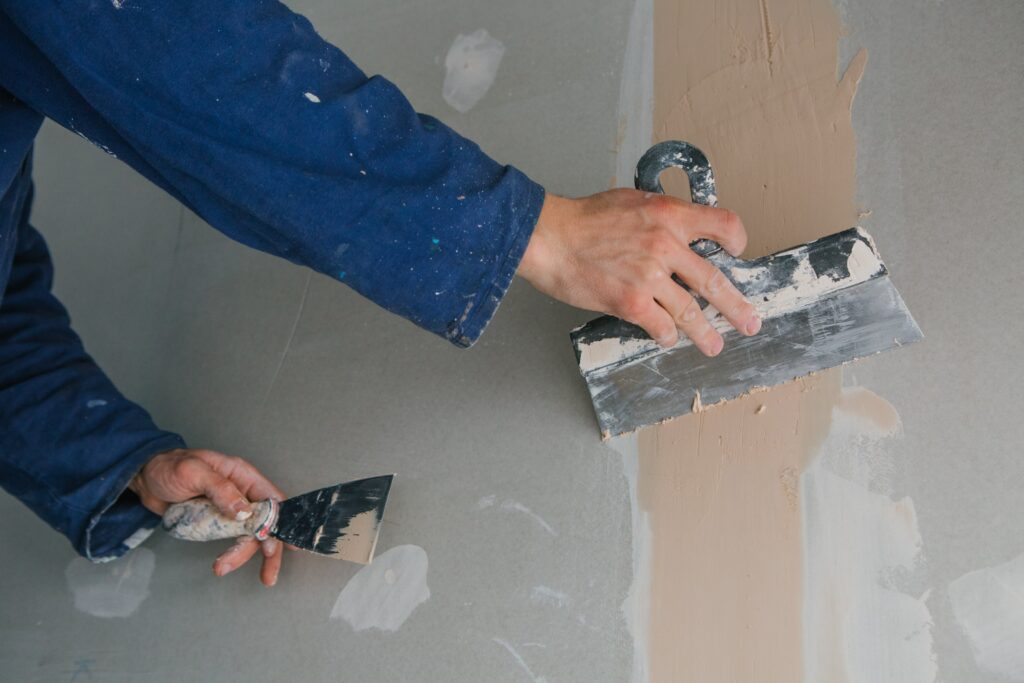 plasterer-man-works-plastering-two-trowels-plasterboard-blue-uniform-min